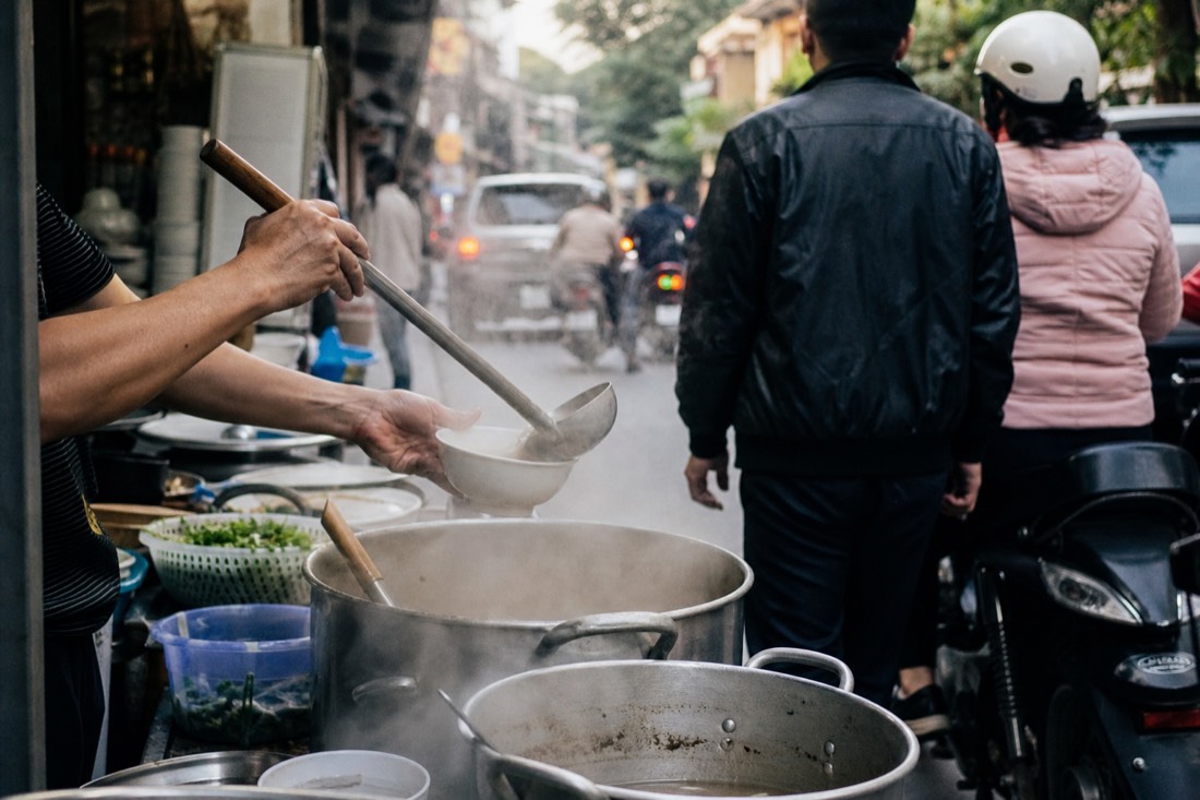 Vendor ladling hot broth from a boiling pot at a busy Hanoi street food stall