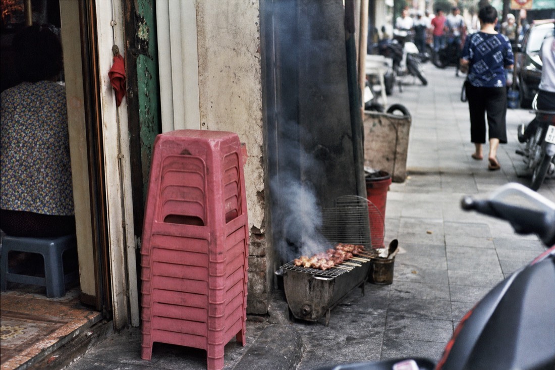 Pavement charcoal grill and stacked plastic stools outside a narrow Hanoi Old Quarter shopfront at midday