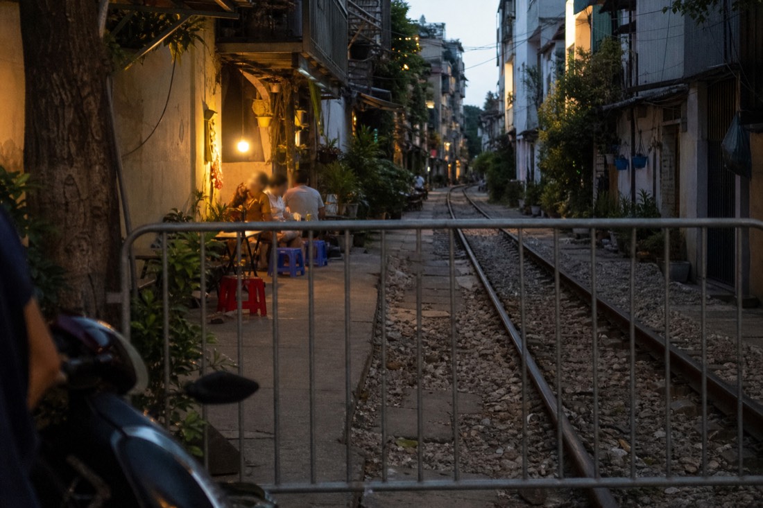 Metal barrier at a Hanoi Train Street entrance with a track-side cafe and stools visible beyond it