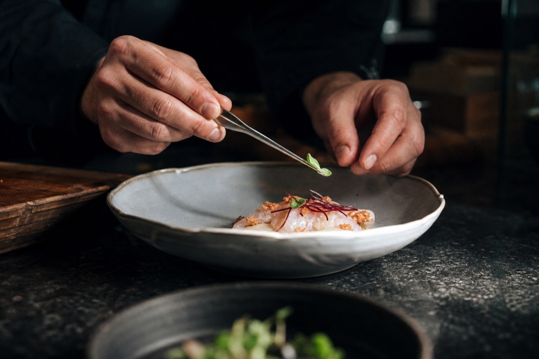 Chef's hands plating a delicate dish with tweezers in a fine-dining kitchen