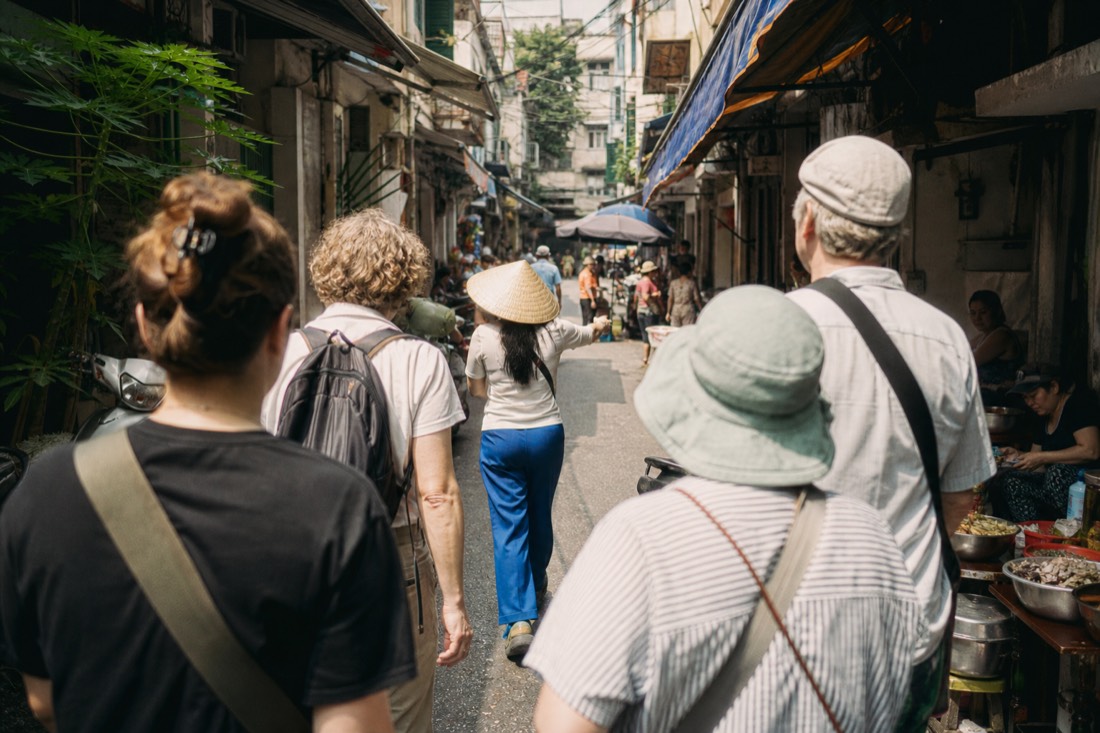 Small group following a local guide past a row of food stalls in a Hanoi lane at midday