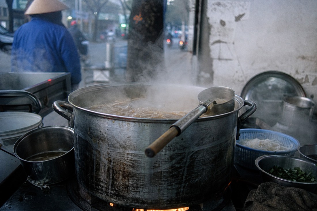 Large metal broth pot simmering over a burner at a Hanoi pho stall at dawn with steam rising