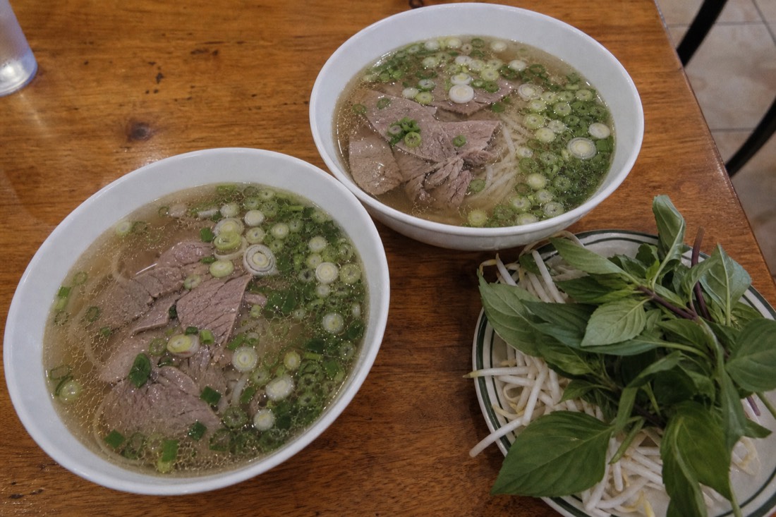 Two bowls of pho side by side on a table, one plain northern style and one with a plate of fresh herbs