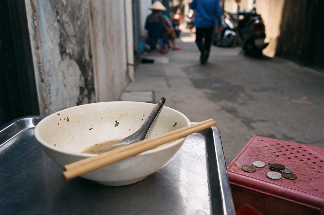 Bowl of pho with a few small coins resting on a low stool beside it at a street stall