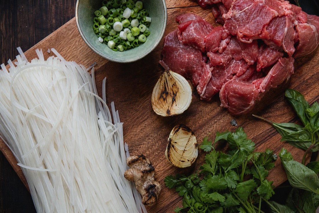 Raw pho ingredients laid out before assembly: rice noodles, sliced beef, charred ginger and onion