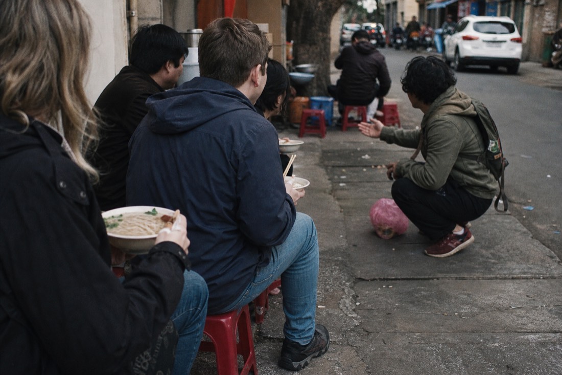 Small group seated on low stools holding bowls of noodles while a guide talks with them at a street stall