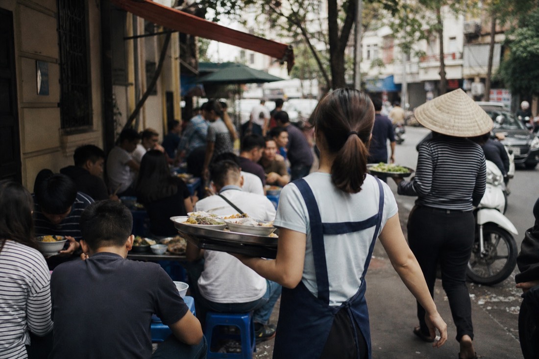 Rows of diners on low plastic stools eating at a busy pavement food stall in Hanoi