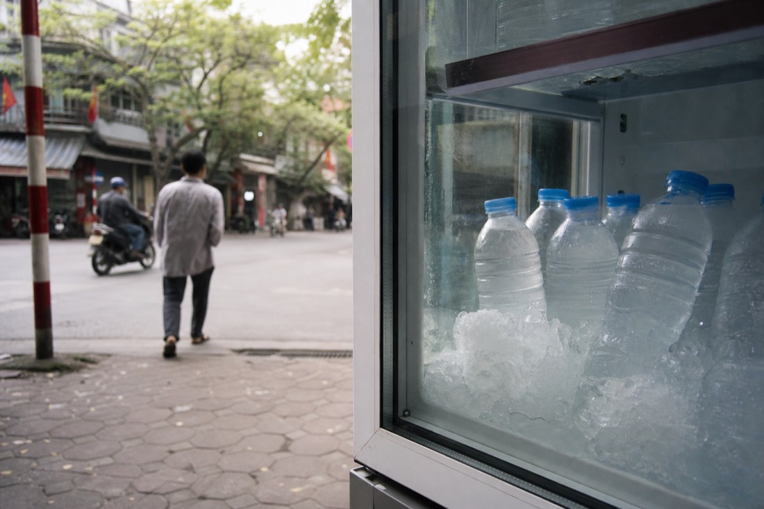 Sealed bottled water on ice inside a glass-front cooler at a Hanoi street corner shop