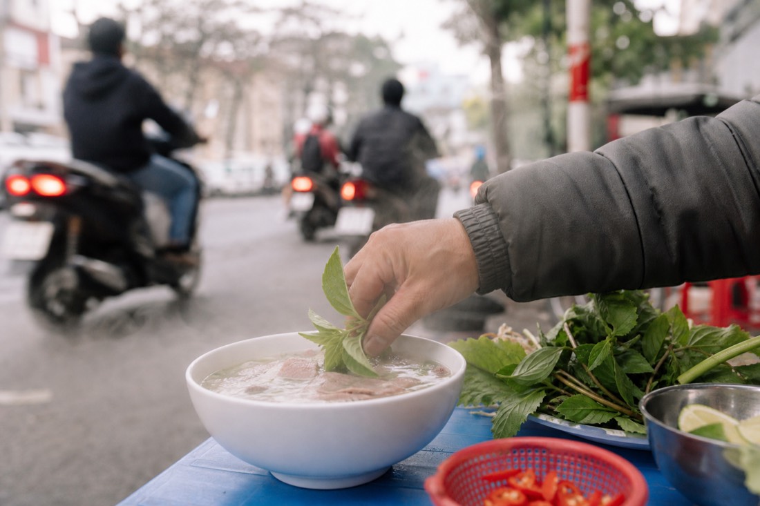 Hand dipping fresh herbs into a steaming bowl of pho broth at a Hanoi street stall
