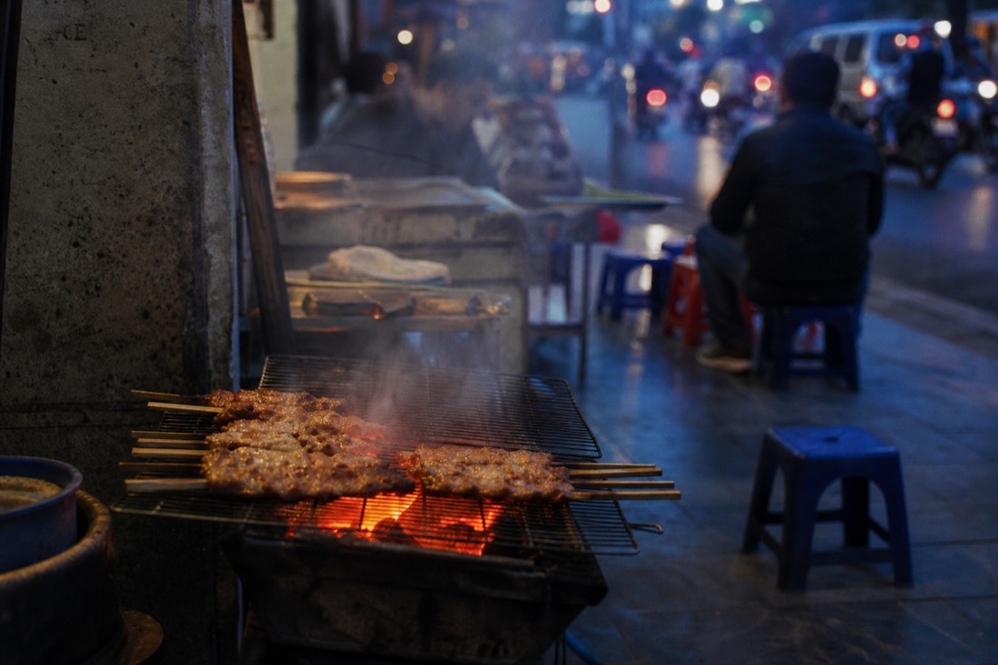 Charcoal grill glowing at dusk with pork skewers cooking at the same Hanoi stall night after night