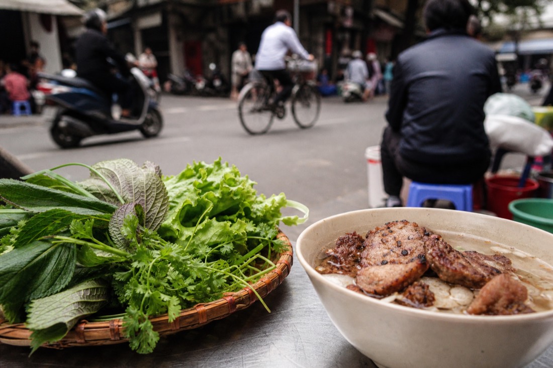 Basket of raw perilla, coriander and lettuce beside a bowl of bun cha at a Hanoi street stall