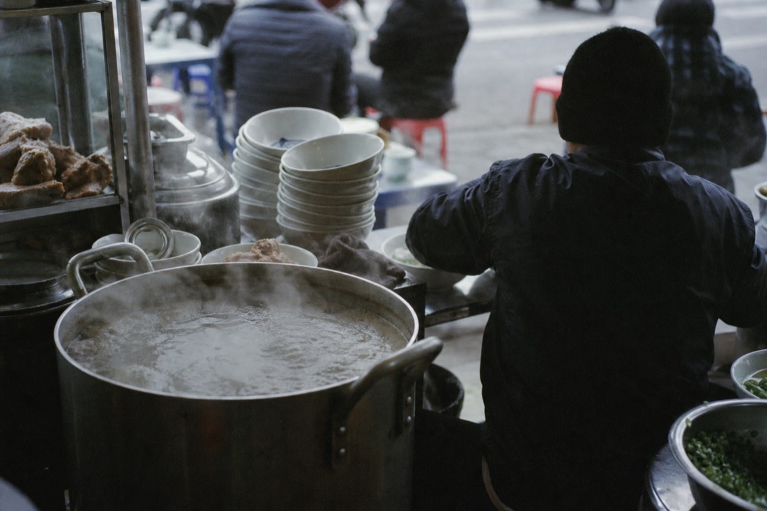 Early morning Hanoi pho counter with broth at a rolling boil and office workers on low stools
