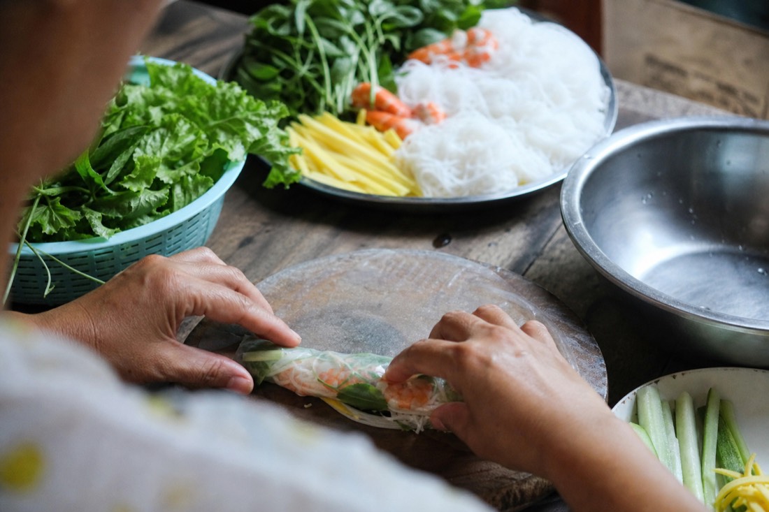 Hands rolling fresh spring rolls in rice paper with vegetables and herbs on a kitchen table