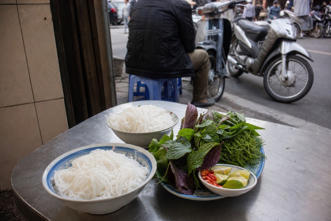 Plate of fresh Vietnamese herbs, lime and chilli beside bowls of noodles at a Hanoi street table