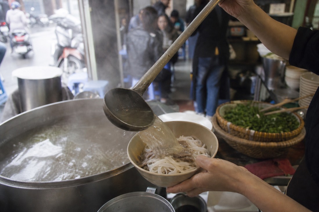 Clear northern-style Hanoi pho broth being ladled over flat rice noodles at a morning shop