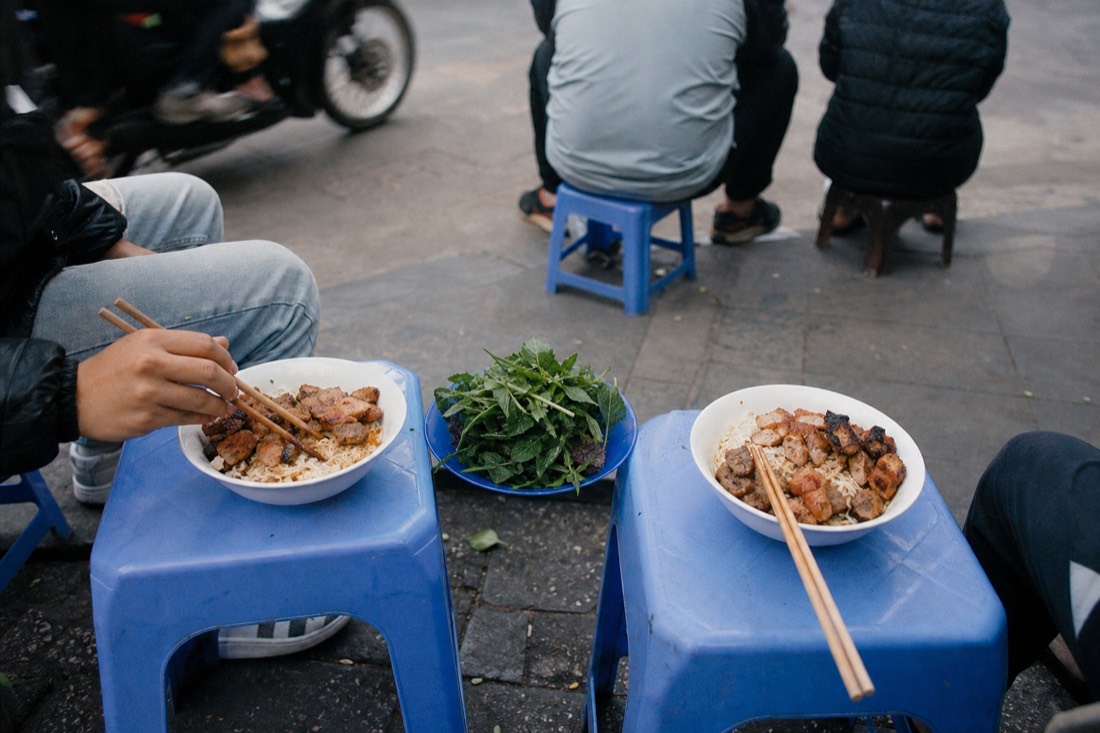 Two people sharing a bun cha set and small dishes at a low table on a Hanoi pavement