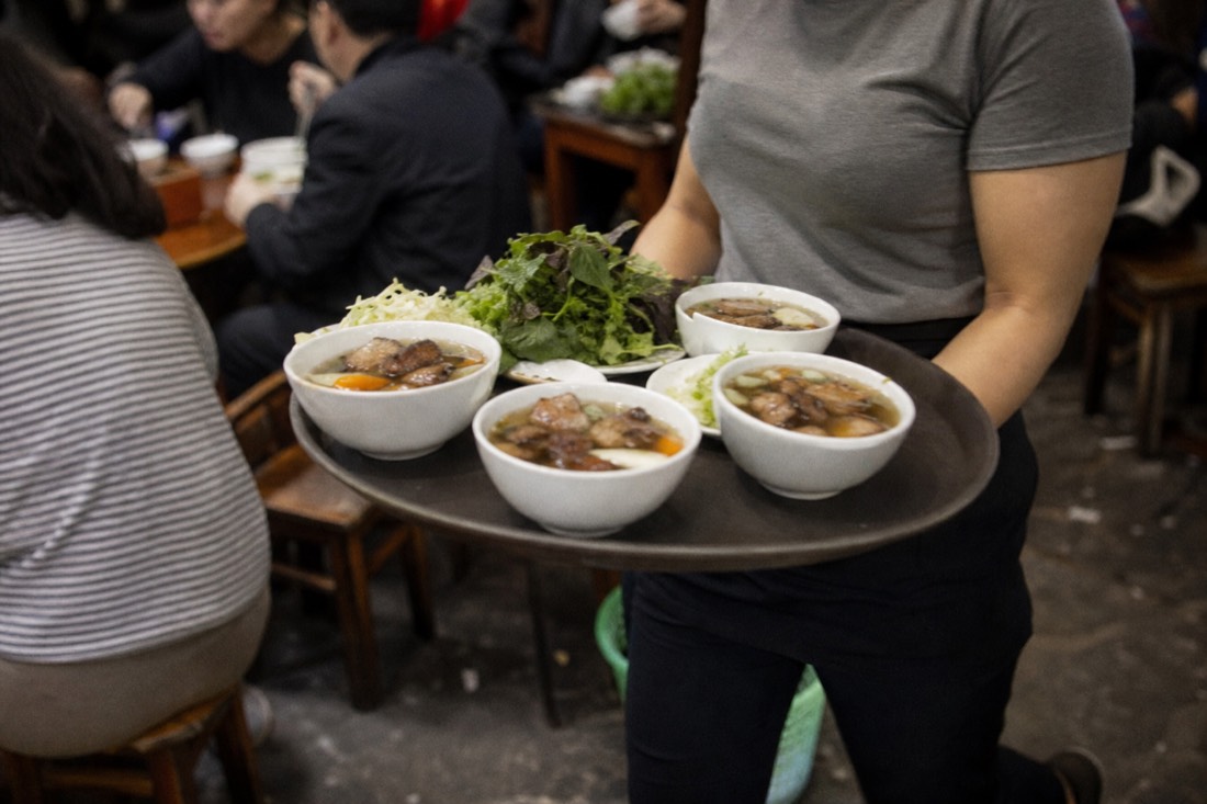 Staff carrying trays of bun cha bowls between crowded tables at a Hanoi restaurant