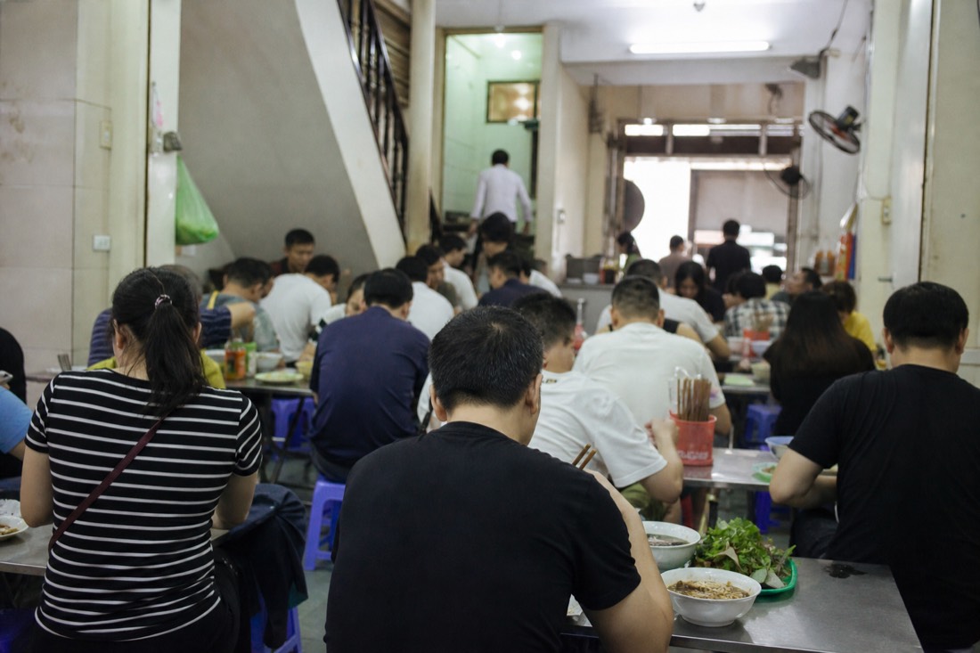 Diners on low plastic stools eating bun cha at crowded tables inside a multi-storey Hanoi restaurant